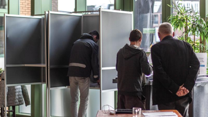 man vult stembiljet in in stemhokje, naast hem deponeren een vrouw en man hun biljet in de bus