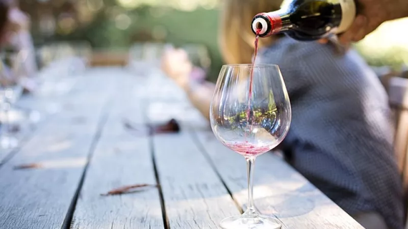 wijnglas op wordt ingeschonken op tafel in tuin, alleen de hand zichtbaar en onscherp vrouw in de achtergrond 
