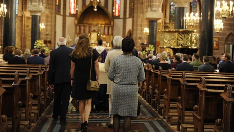 nabestaanden (op de rug gezien) volgen grafkist tijdens begrafenisdienst, links en rechts kerkbanken met mensen, voorop loopt een priester
