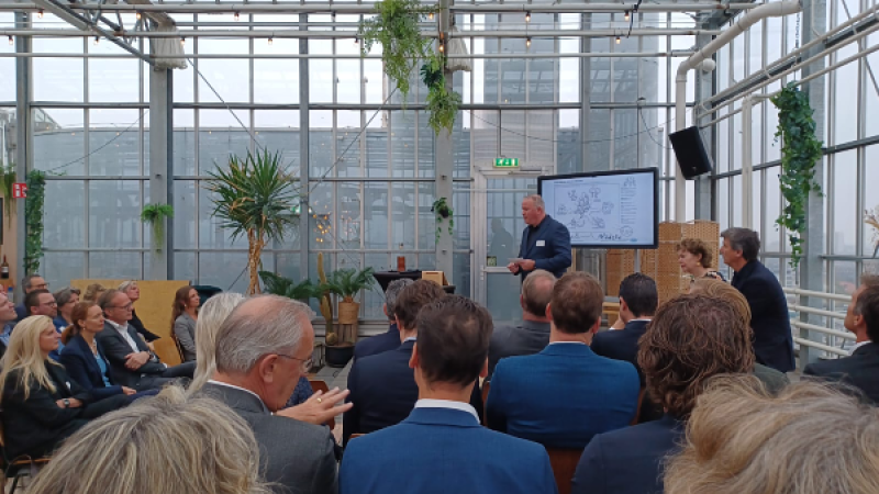eerste rijen deelnemers (deels op de rug, deel van opzij) terwijl Frank den Brok het woord voert staande voor een scherm met afbeelding