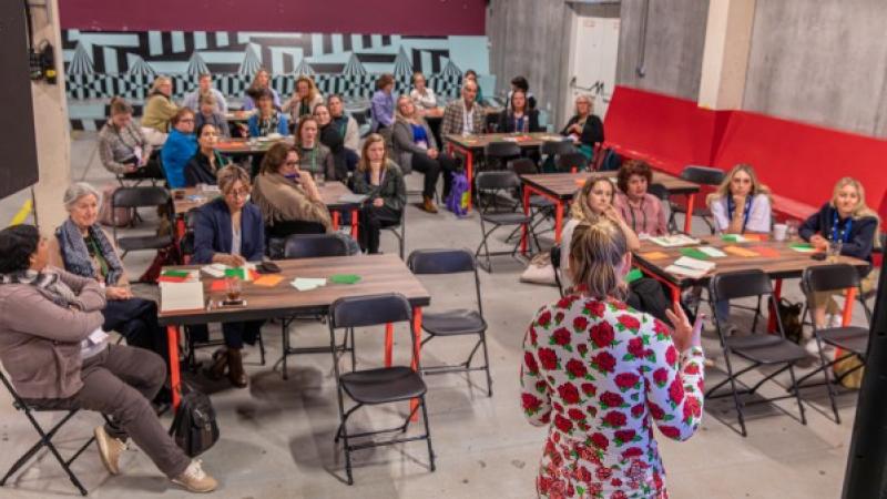 Een groep mensen aan tafels luistert naar een docent