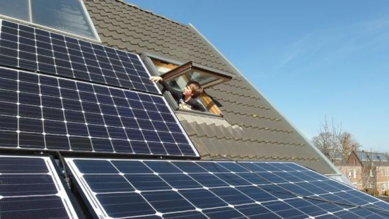 Een jongen steekt vanuit een kantelraam zijn hand uit naar de zonnepanelen op het dak