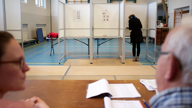 vrouw brengt stem uit in stemlokaal, stembureauleden kijken toe
