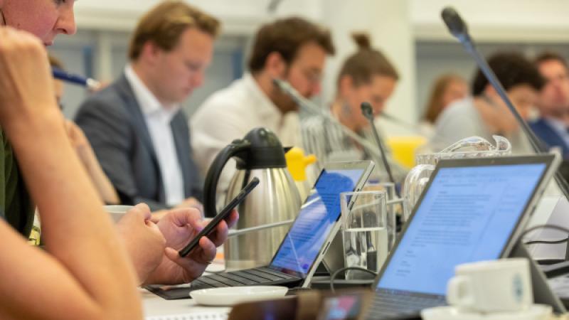 Raadsleden aan een vergadertafel met onder meer laptops en een koffiekan