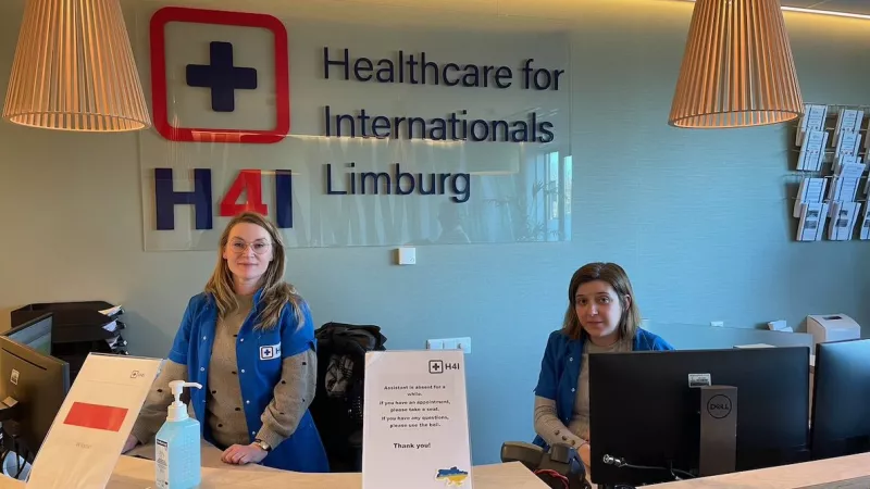 Twee vrouwen staan achter de balie van de huisartsenpraktijk ‘Healtcare for Internationals Limburg’ 