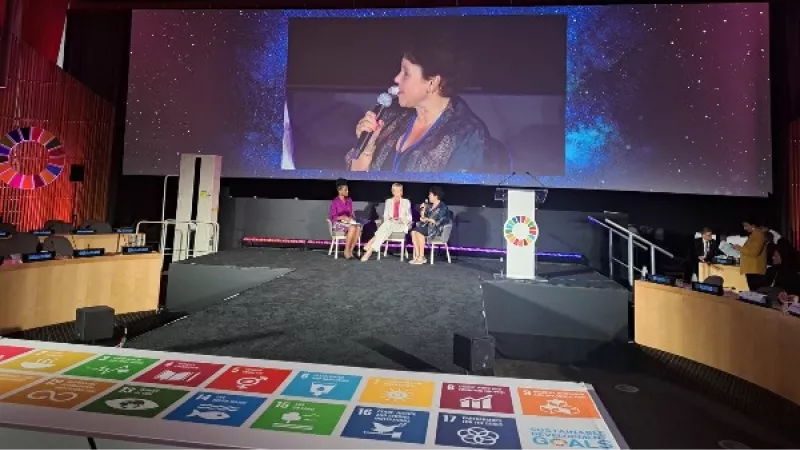 Sharon Dijksma in gesprek met 2 andere vrouwen, zij wordt getoond op een groot scherm, met afbeelden van de Global Goals op de voorgrond