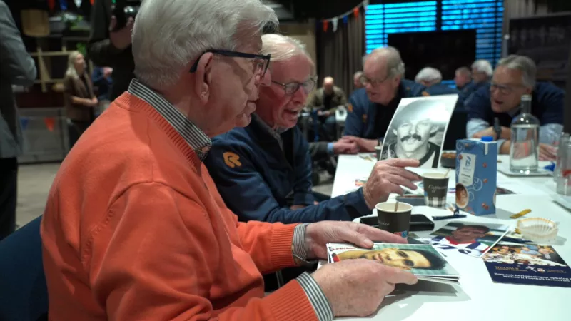 twee oudere mannen bekijken foto's aan een tafel met daarop koffiebekers