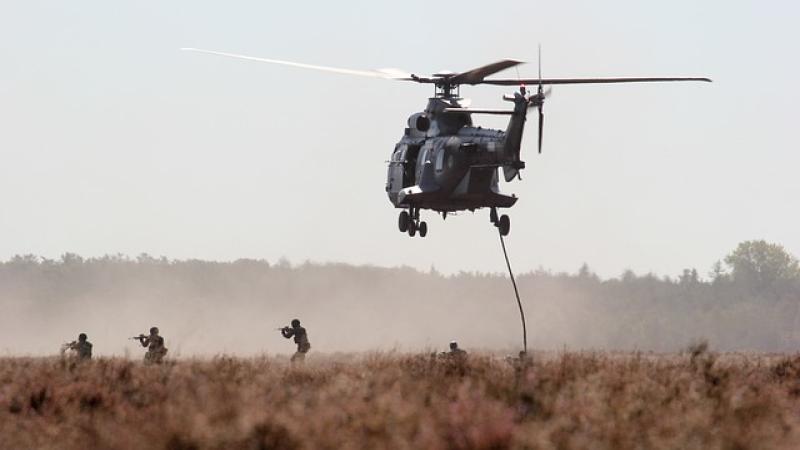 soldaten in met geweren in actie op heide-achtig terrein, boven hen een laagvliegende helikopter