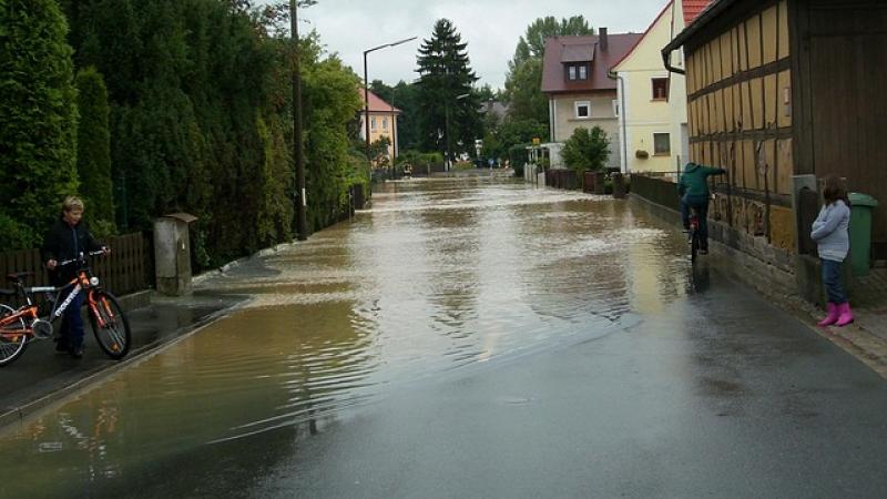 Overstroomde straat met aan de ene kant bomen en aan de andere kant huizen, een fietser (op de rug gezien) rijdt door het water, links staat een jongetje met zijn fiets, rechts staat een meisje die de kijkt naar de fietser.