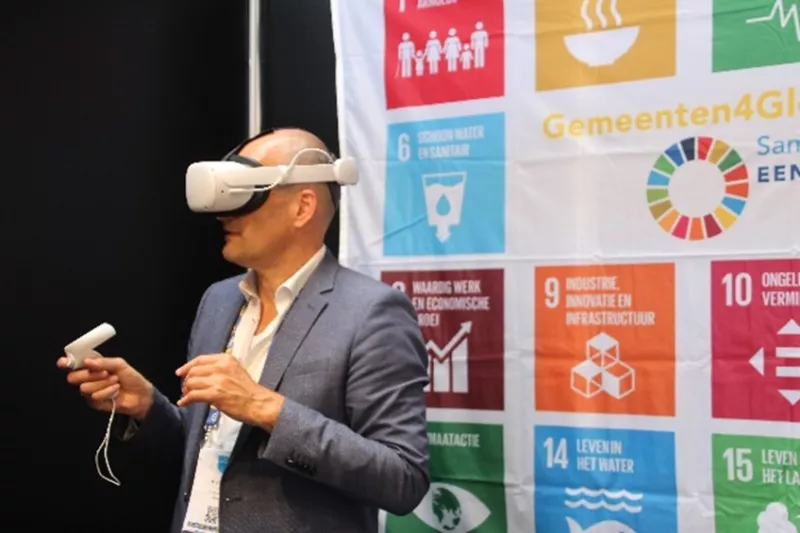 Man met een VR-bril op en een controller in zijn hand, voor een vlag van Gemeenten4GlobalGoals