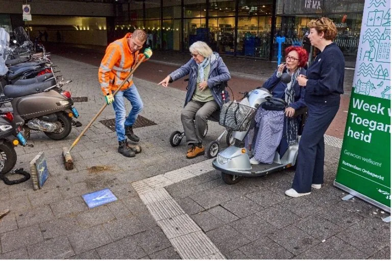 Arja zittend in een scootmobiel op straat in Rotterdam. Naast haar staat een mevrouw, aan de andere kant zit een mevrouw op haar rollator, ze is in gesprek met een straatveger. Rechts staat een bord met de tekst 'Week van de Toegankelijkheid'