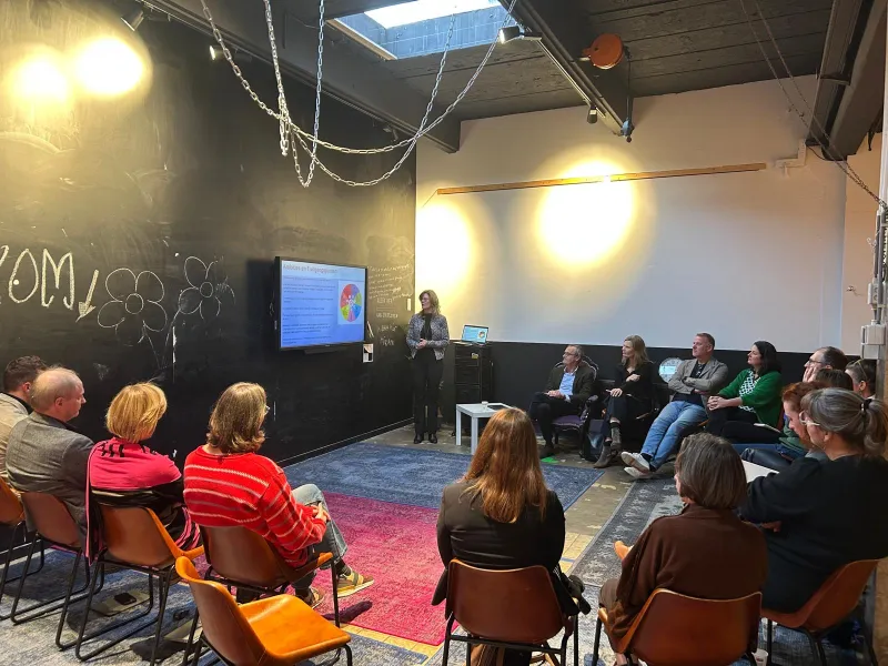 deelnemers zitten in een kring en luisteren en kijken naar een presentatie op een scherm waar een vrouw bijstaat
